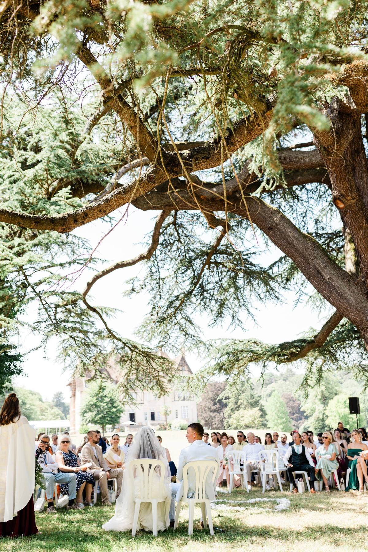 Outdoor wedding ceremony under gnarled tree with guests seated facing bride and groom near historic church building.