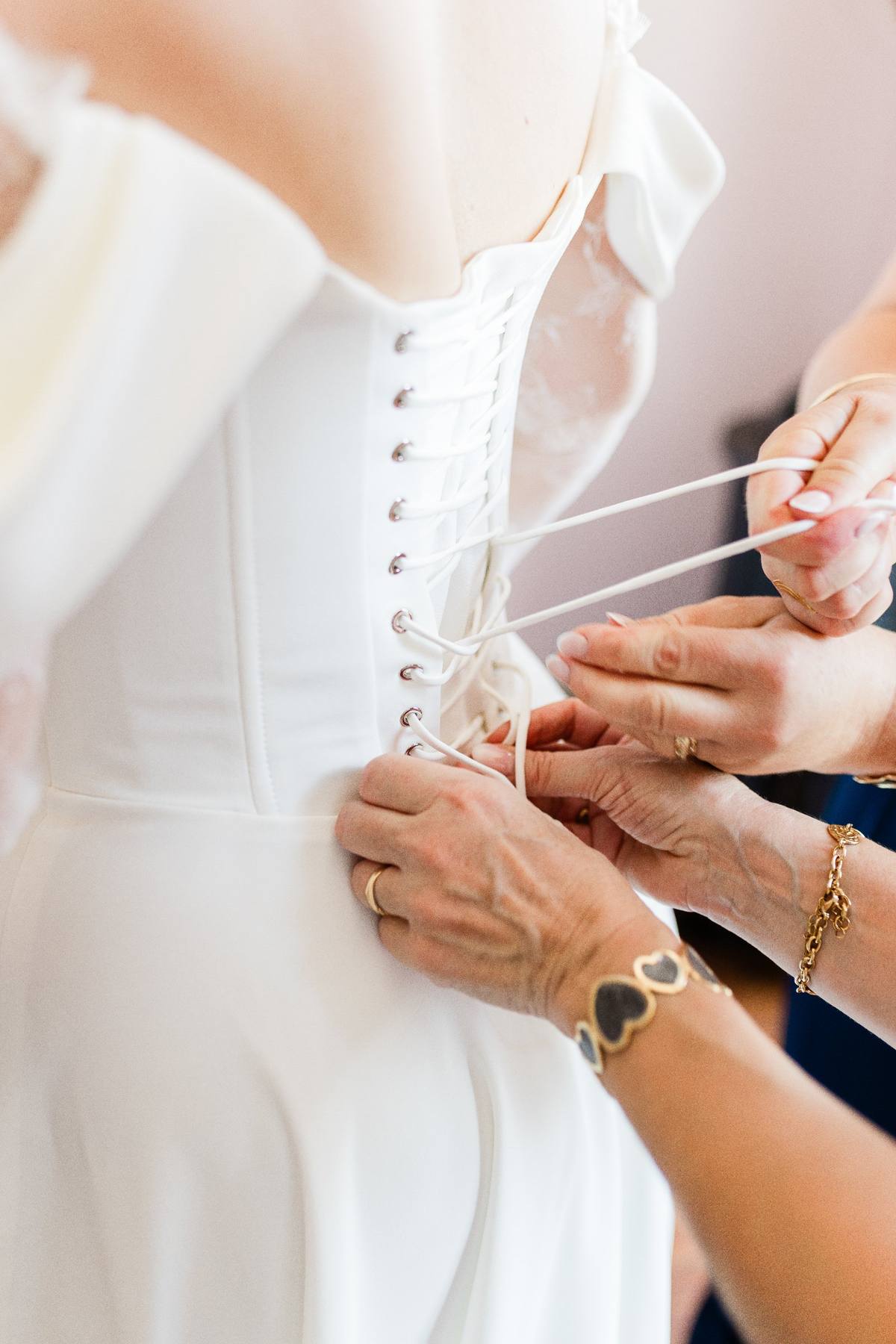 Bride in white dress having corset laced by bridesmaids wearing gold bracelets and rings
