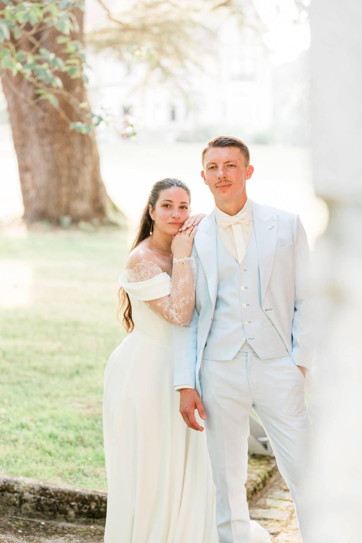 Bride and groom in light clothing posing together beneath a large tree outdoors