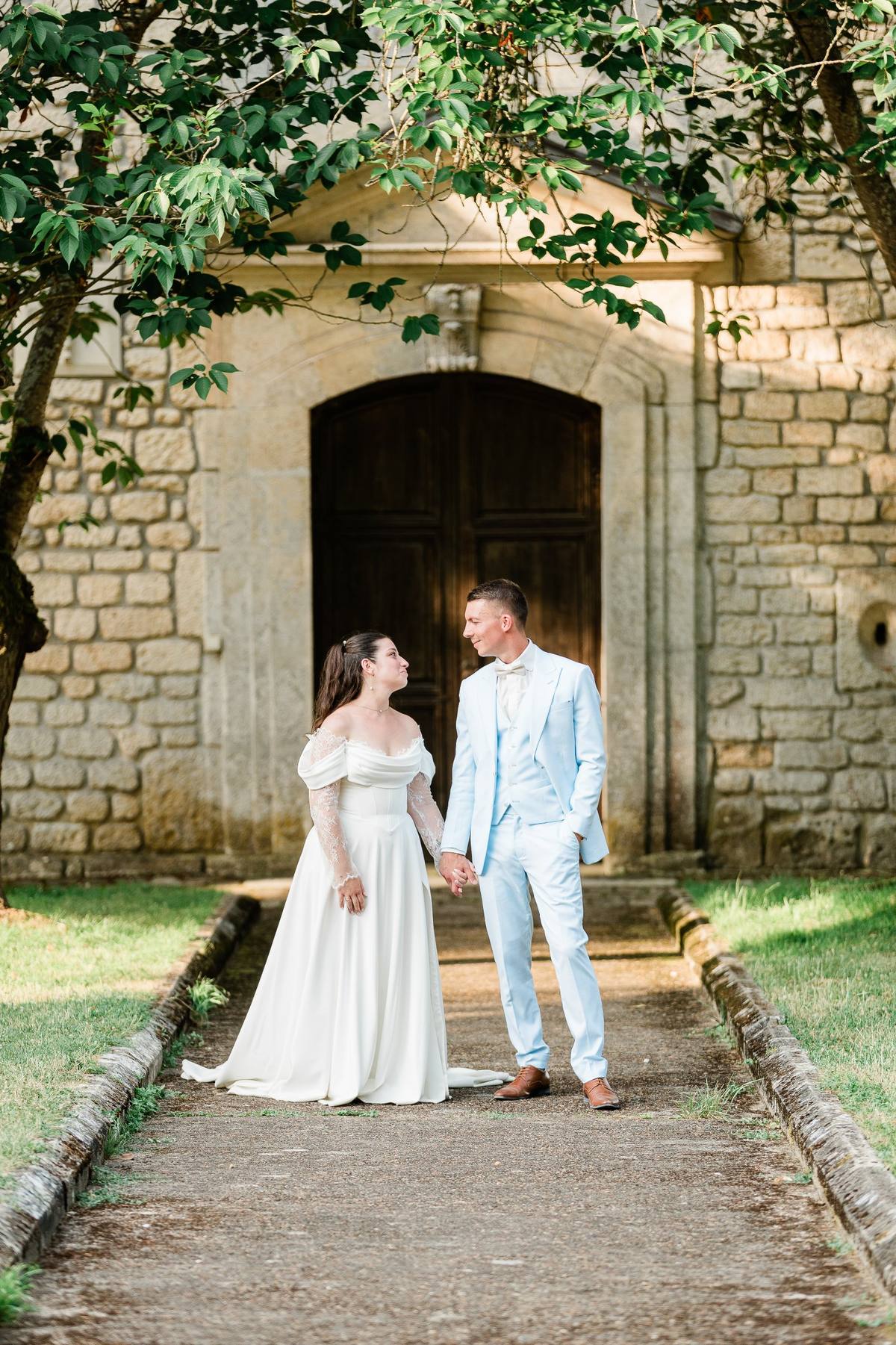 Un couple de mariés se tient la main devant une église en pierre ancienne, sous des branches de lierre.
