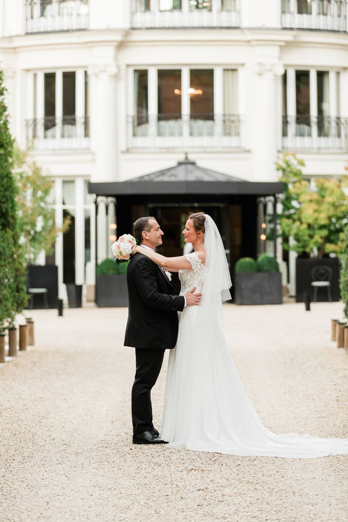 Un couple de mariés pose devant un bâtiment blanc élégant dans une cour pavée.