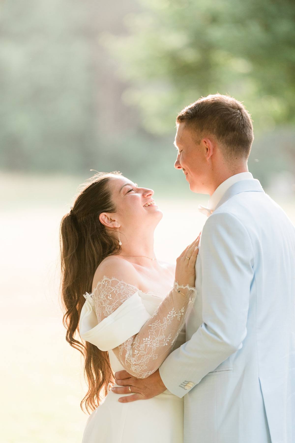 Un couple en tenue de mariage sourit en se regardant dans un jardin ensoleillé.
