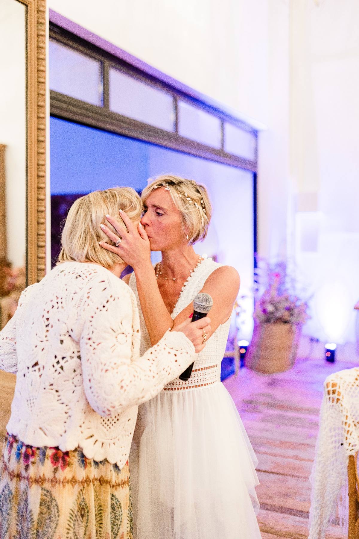 Deux femmes en blanc s'embrassent lors d'une cérémonie avec éclairage violet ambiant.