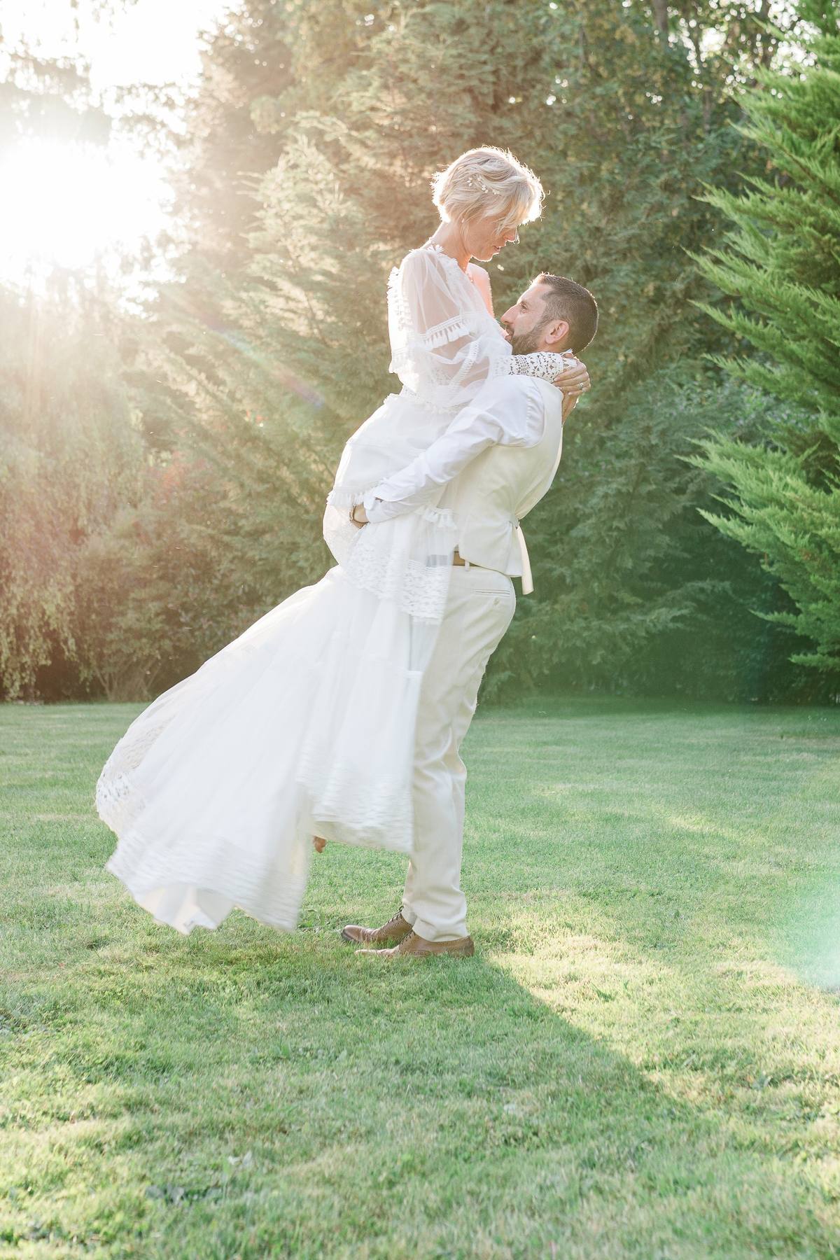 Un homme en costume clair soulève une femme en robe blanche dans un jardin ensoleillé.