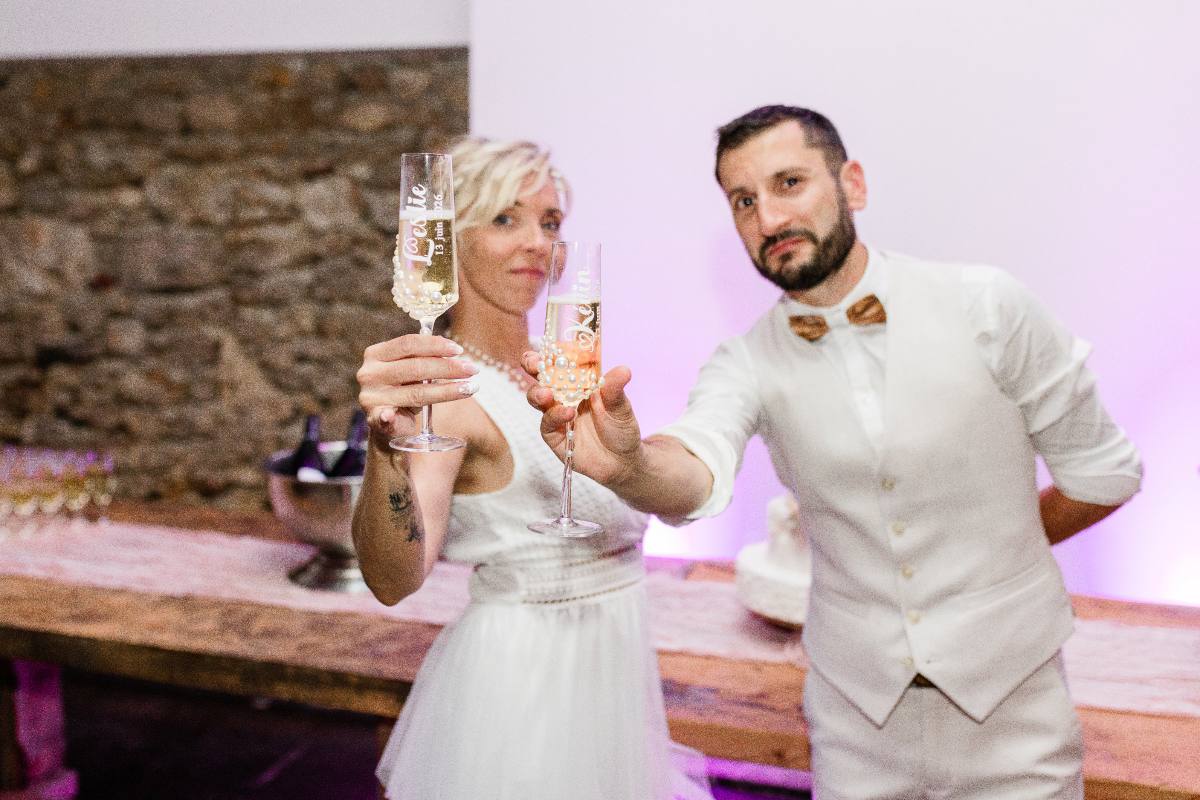 Un couple en tenue blanche lève des verres de champagne dans un décor de pierre