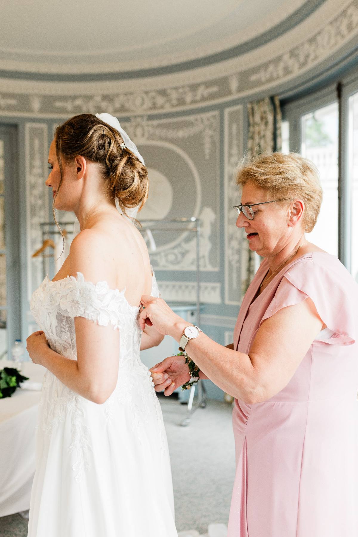 Une femme en robe blanche de mariage ajuste sa tenue avec une femme plus âgée en rose dans une salle élégante.