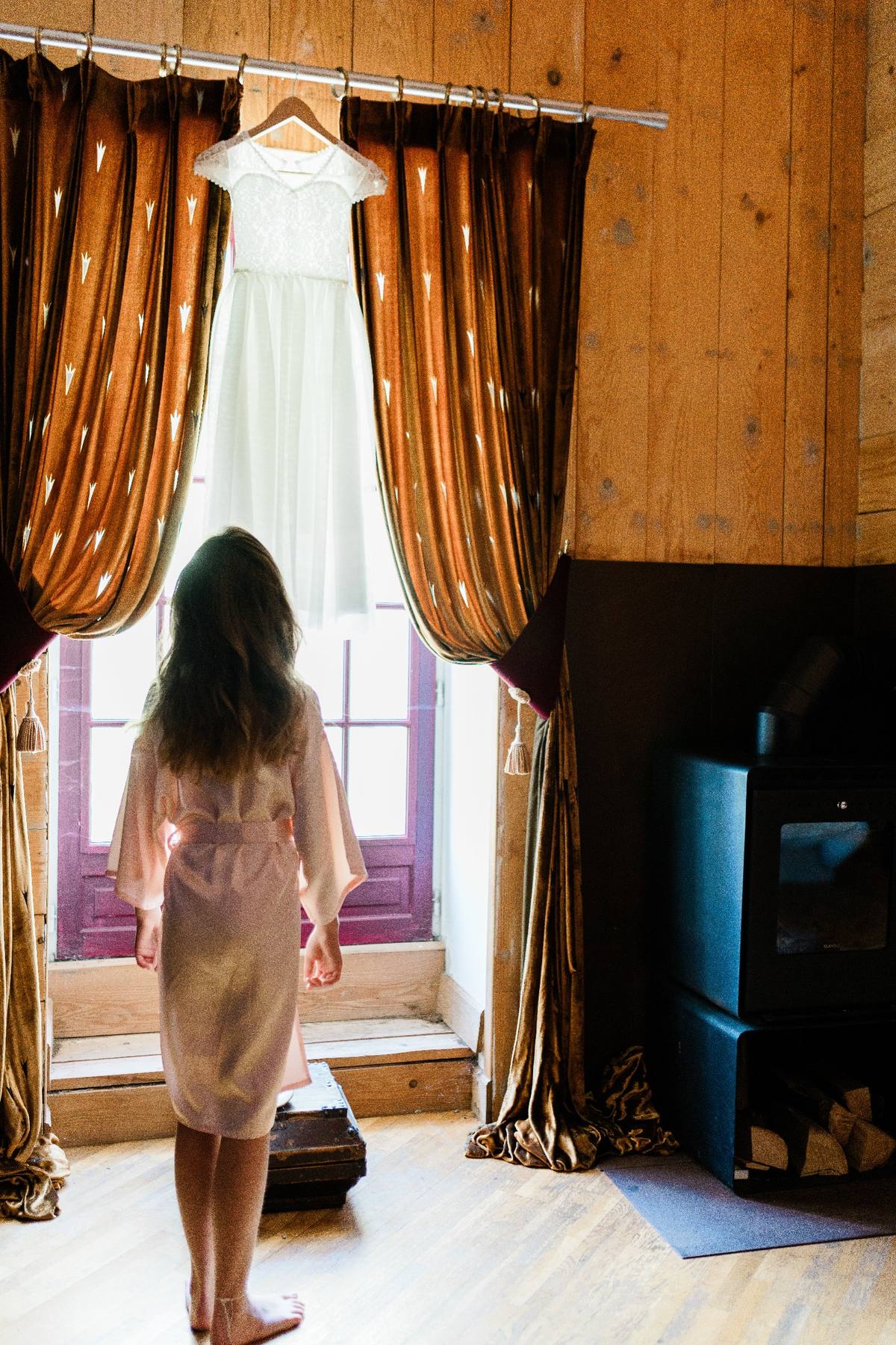 Une femme de dos regarde une robe blanche suspendue, entourée de rideaux dorés dans une pièce en bois.