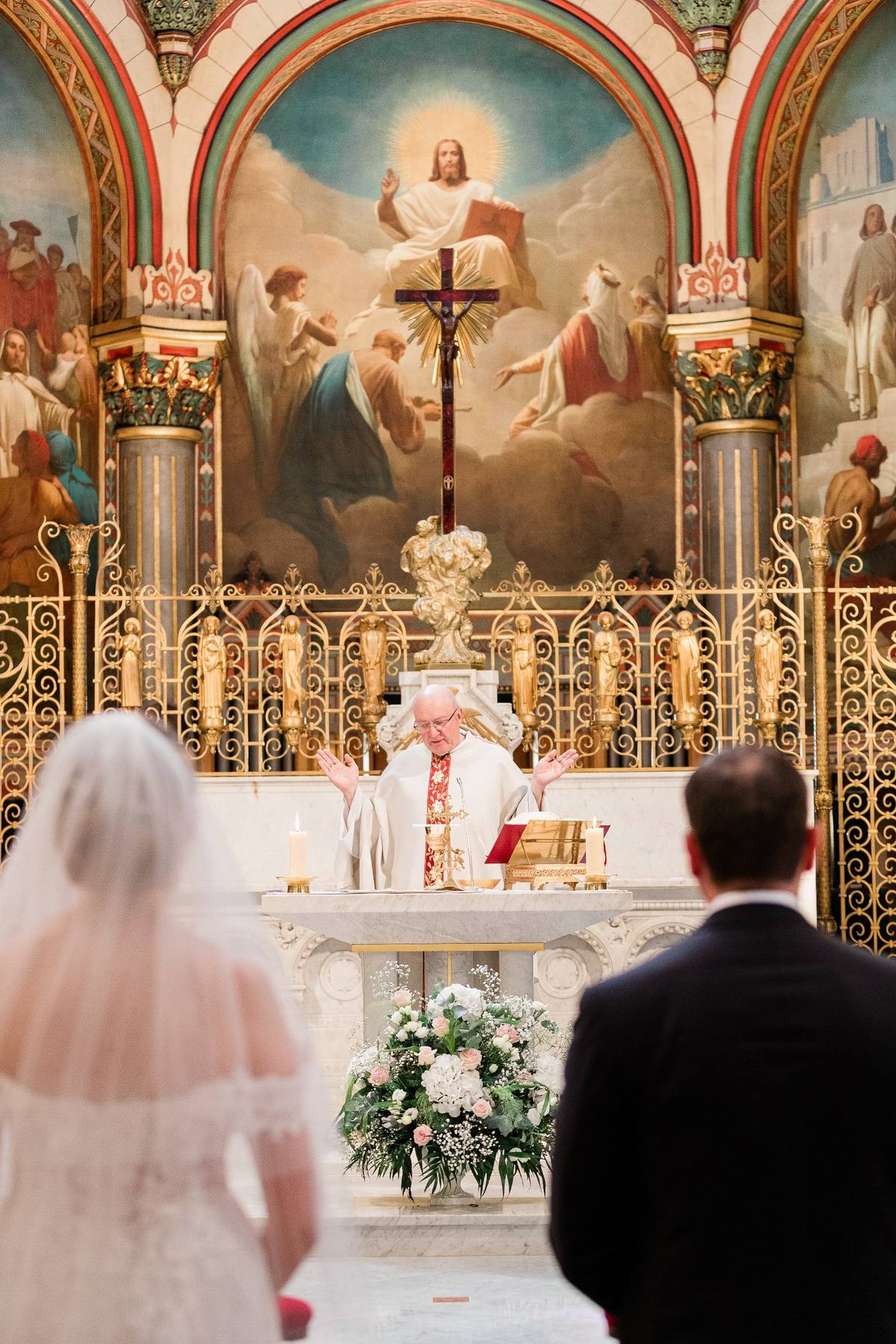 Prêtre célébrant la messe devant un autel orné, couple de mariés en premier plan, église décorée richement.