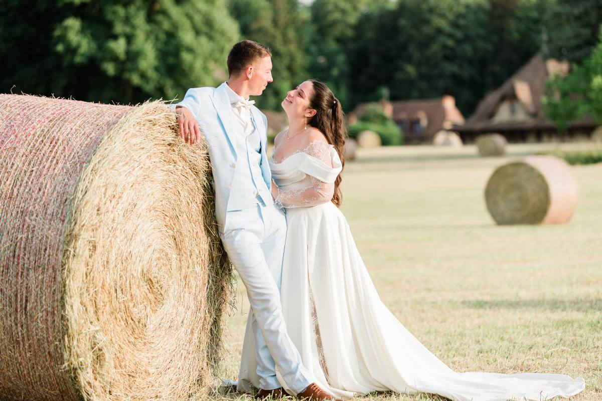 Bride and groom in formal wear posing beside a large hay bale in a rural field.