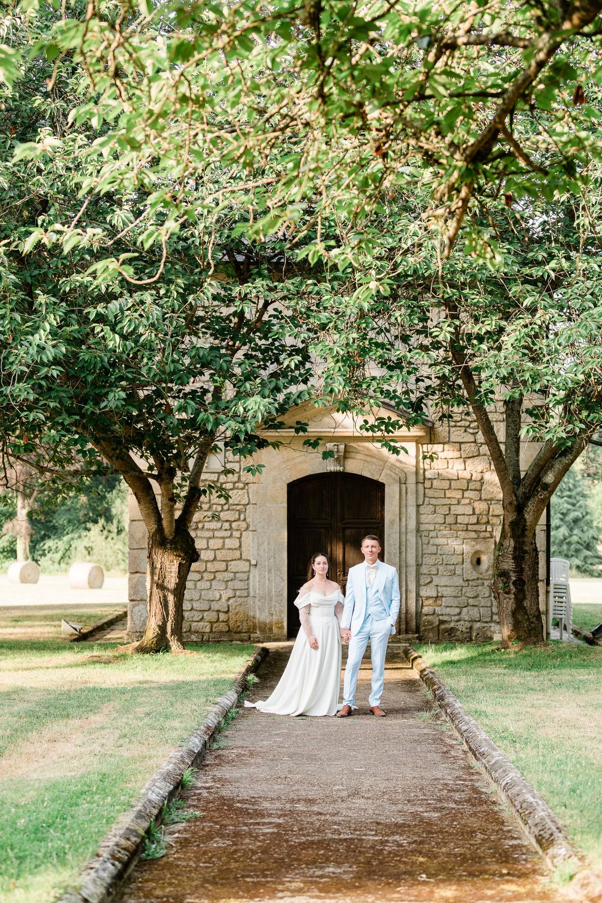 Bride and groom stand on a tree-lined path before a stone building with dark doors.