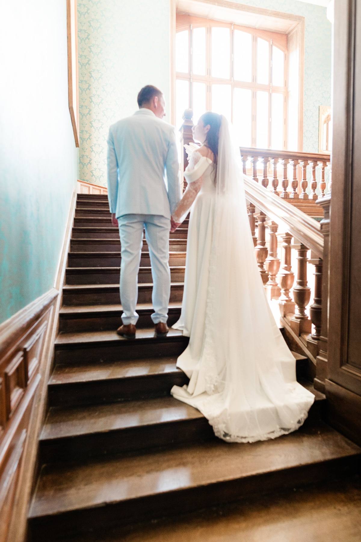 Bride and groom holding hands on wooden staircase in bright sunlit hallway.