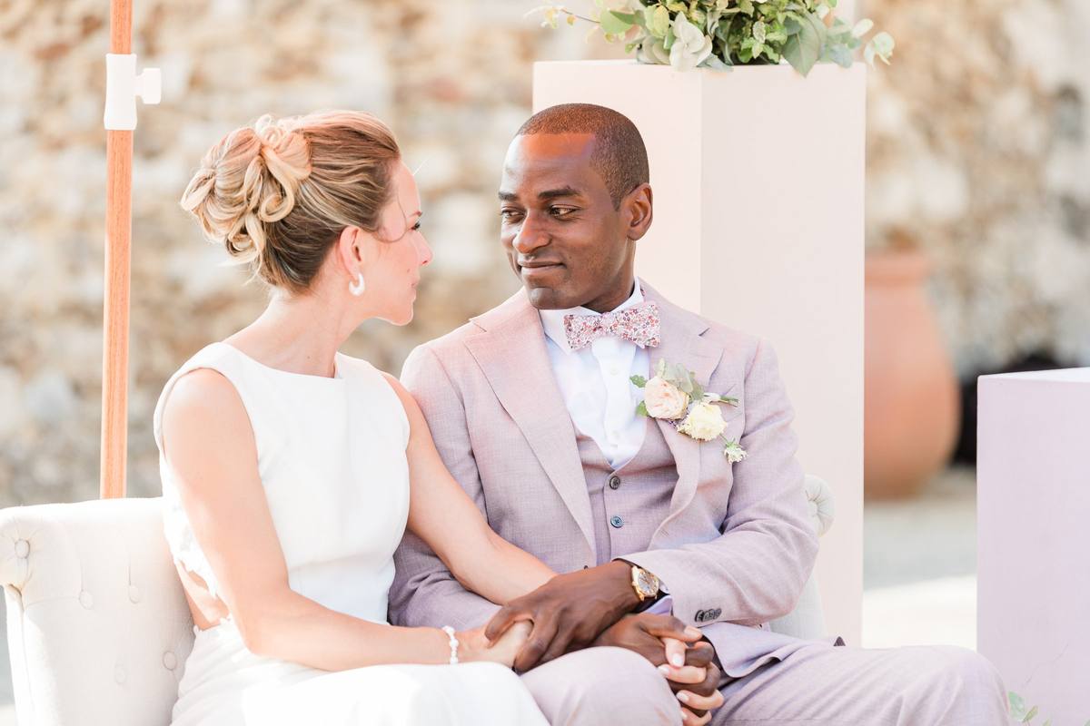 Bride and groom sit together smiling at outdoor wedding ceremony with rustic brick backdrop.