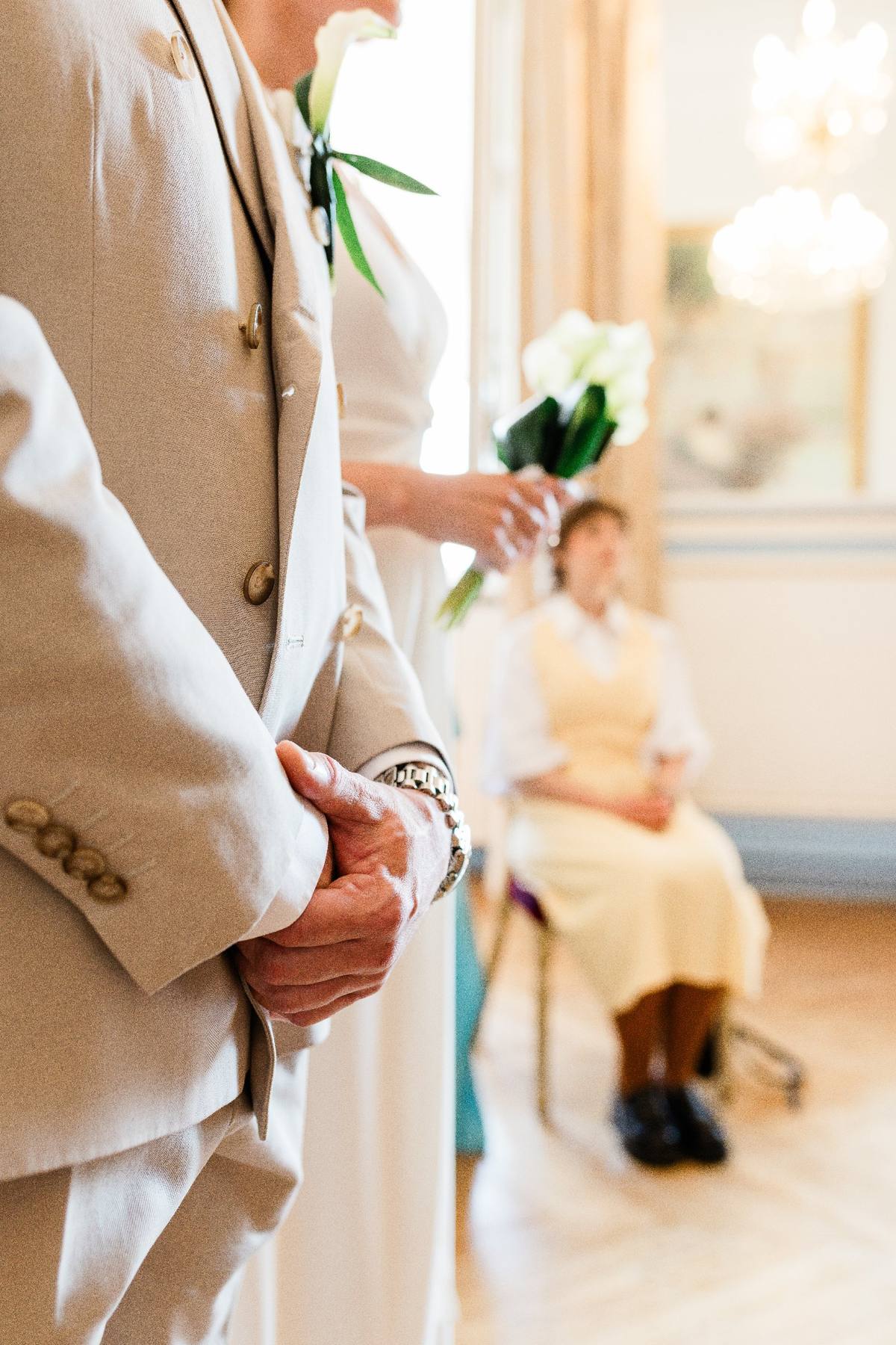 Groom in beige suit holding white flowers with wedding ring visible, bright natural light from window