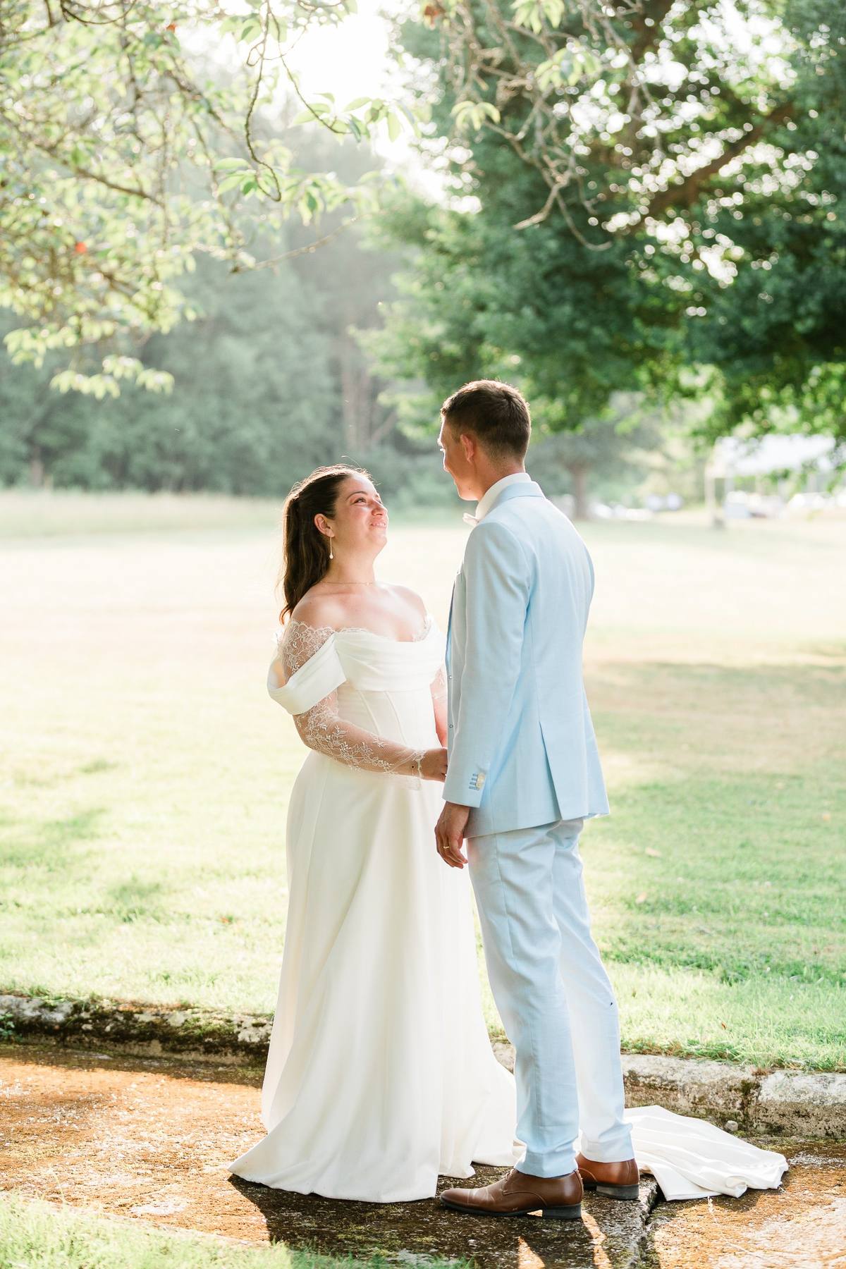 Bride and groom stand together under tree canopy in sunlit park setting.