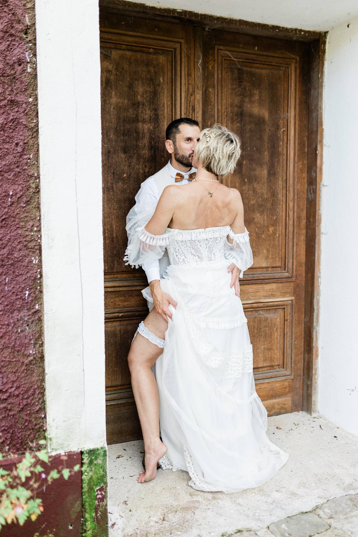 A couple in wedding attire stands intimately by a weathered wooden door in an old stone alcove.