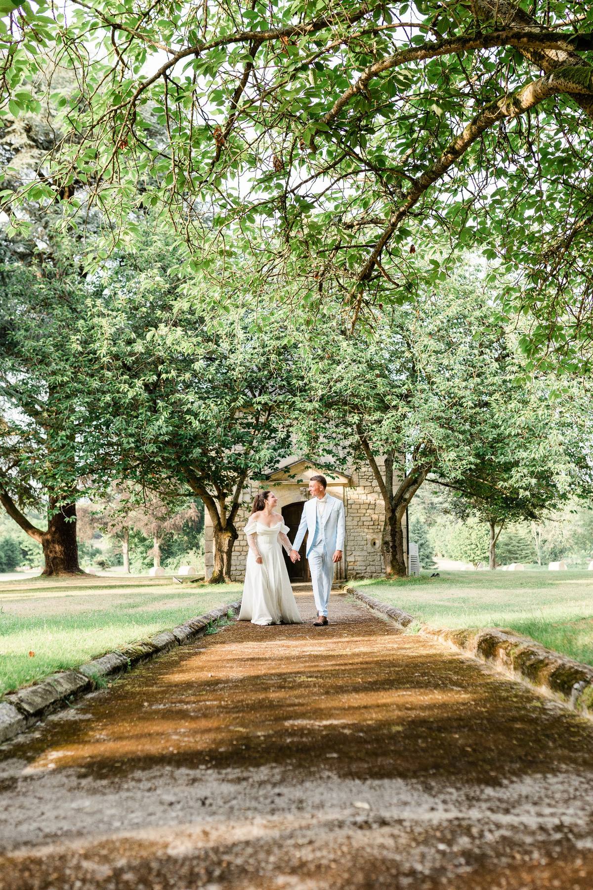 Bride and groom holding hands on tree-lined path toward stone chapel