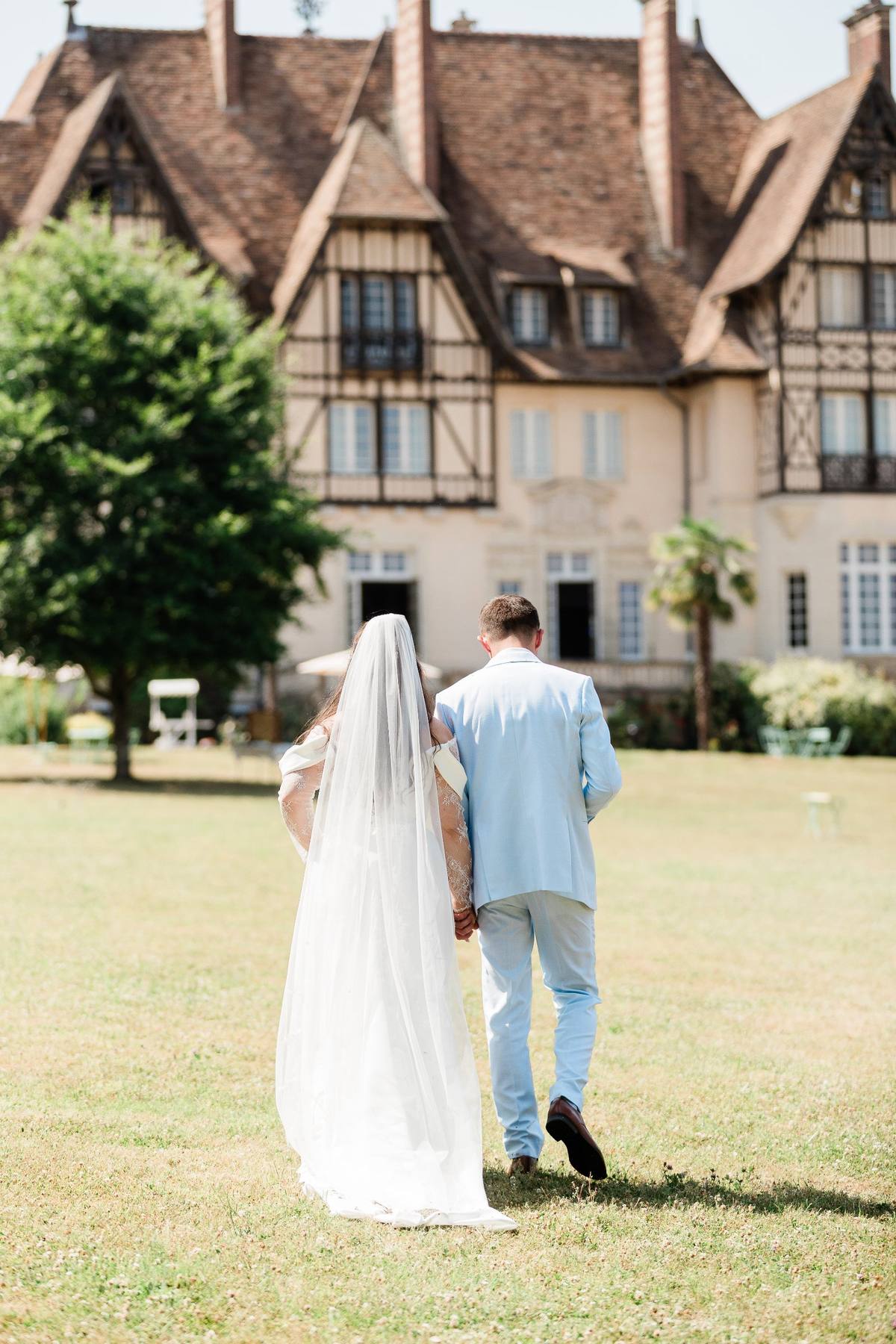Bride and groom walking away from half-timbered manor house on sunny lawn