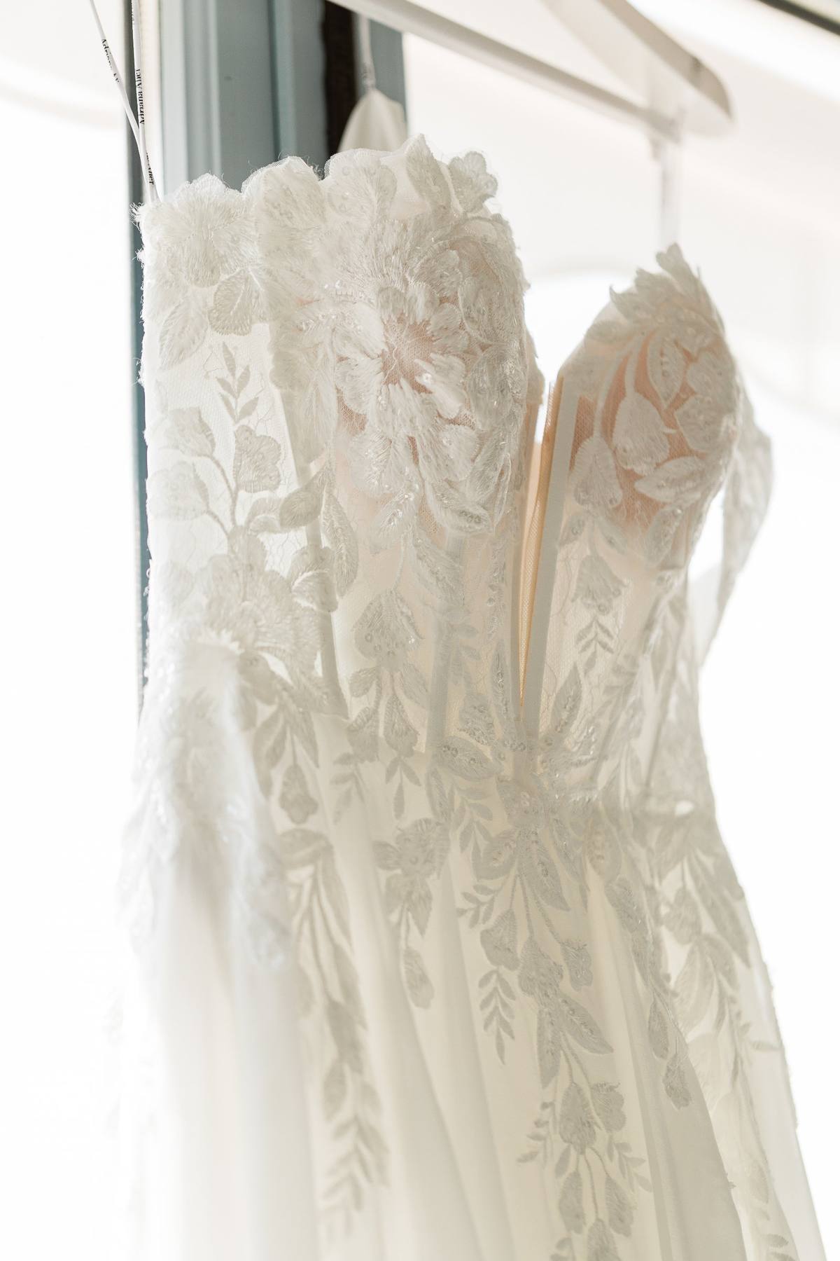 Delicate white lace wedding dress hanging by window in natural light