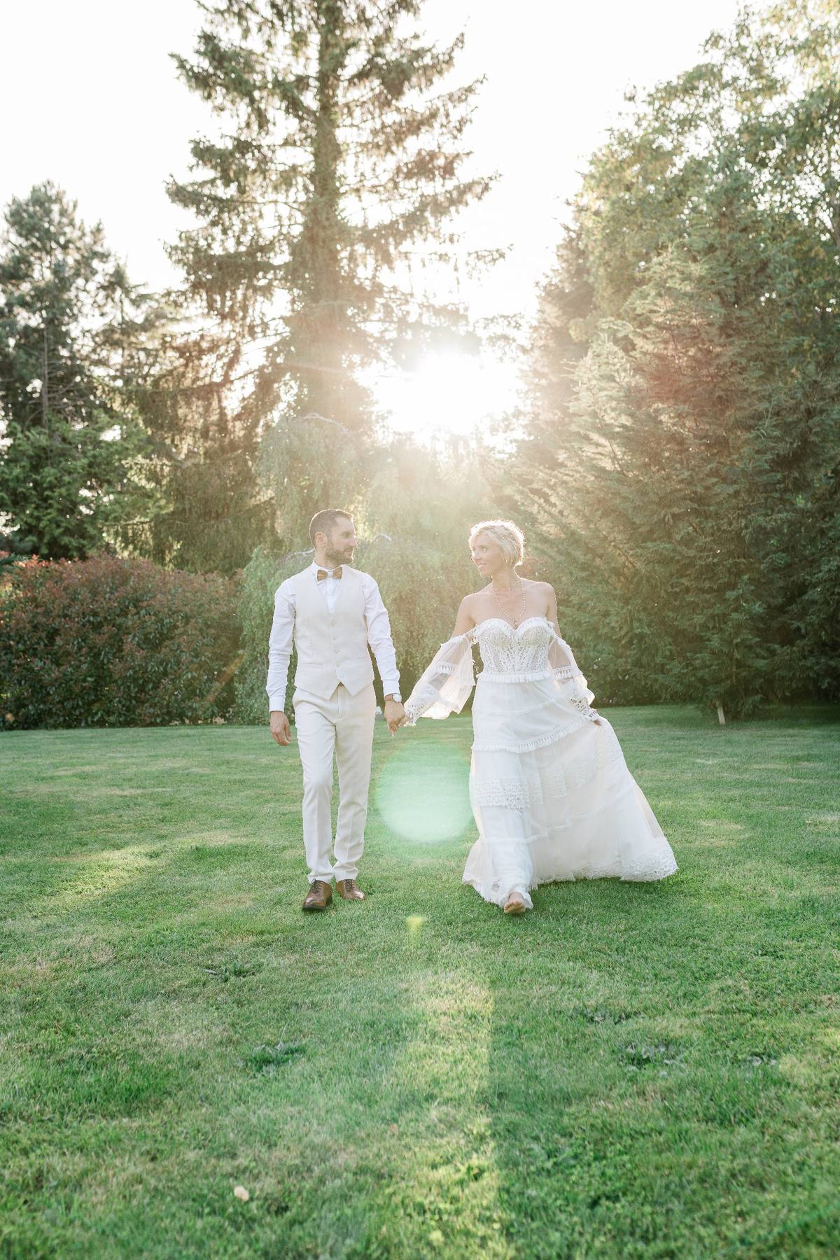 Newlyweds in white attire holding hands walking through sunlit garden with tall trees.