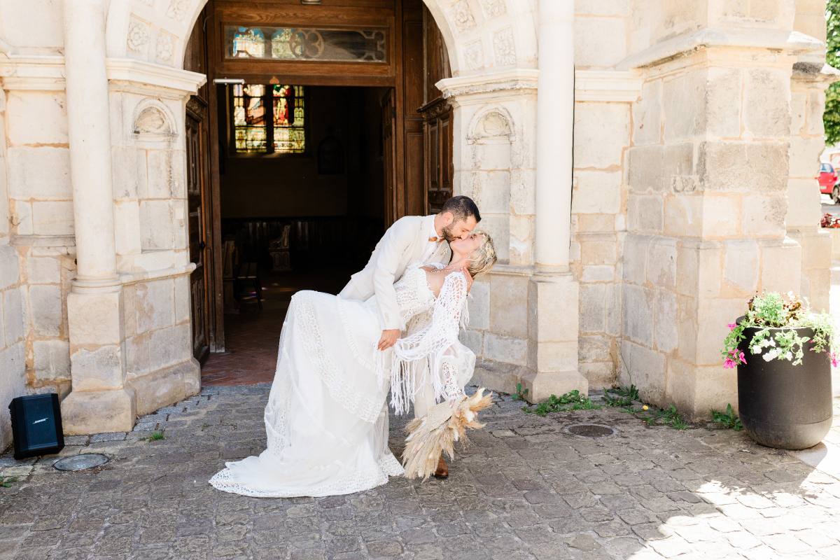 Groom dips bride in white lace dress outside stone church entrance with pampas grass bouquet