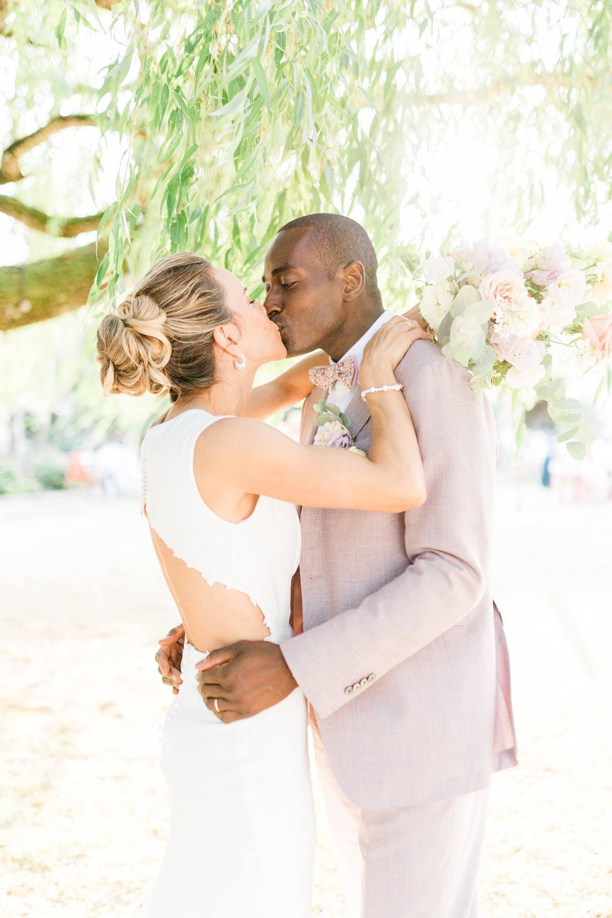 Newlyweds kissing under hanging willow branches with flowers on a sandy beach during daytime