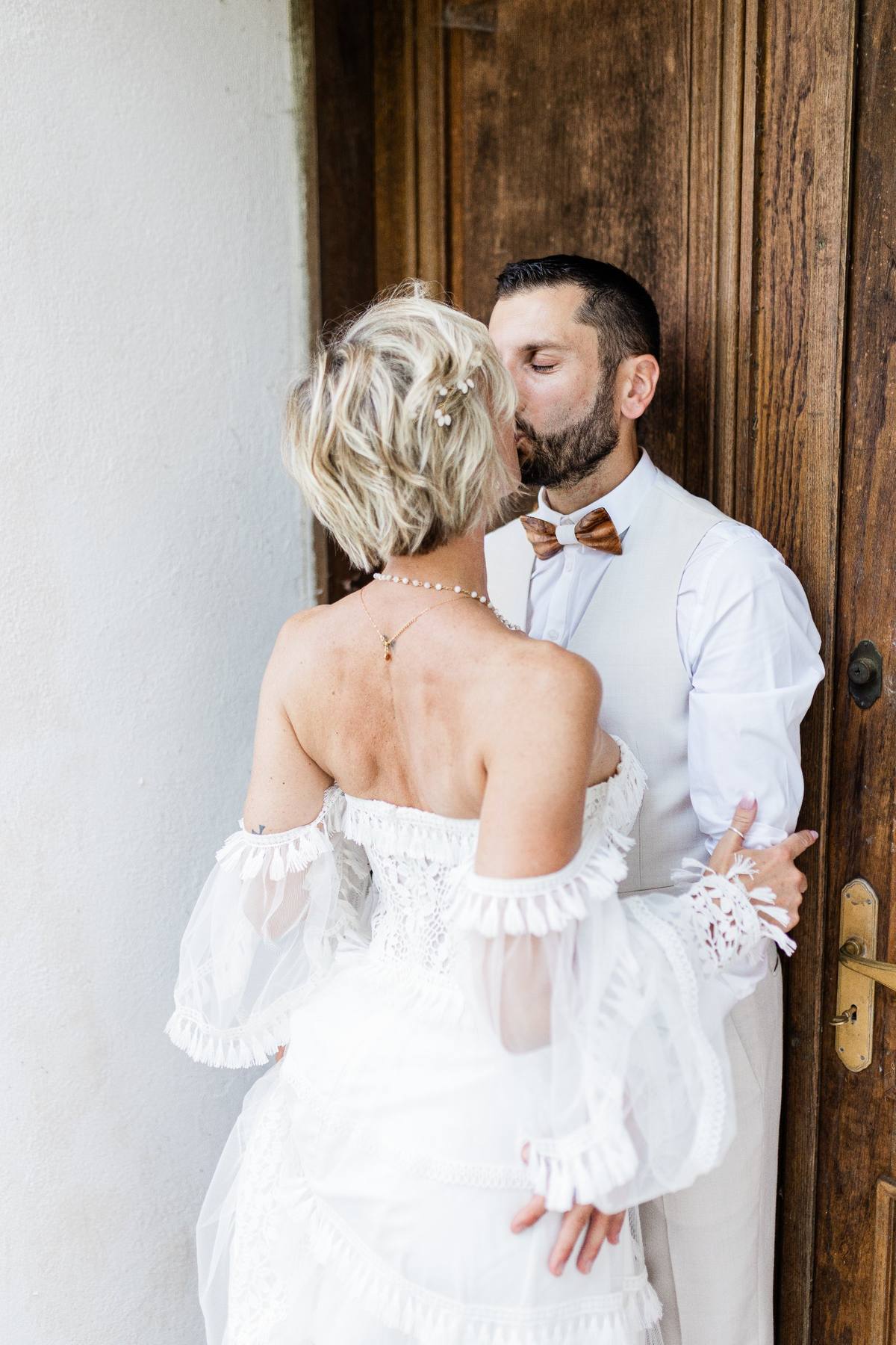 Couple in formal wedding attire embracing against a wooden door and white wall.